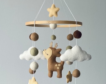 Peut inclure: Un cerf en feutre marron avec des yeux blancs et des bois en paillettes dorées est suspendu à un anneau en bois avec des nuages en feutre blanc, des étoiles et des pompons marrons et verts.