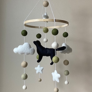 May include: A wooden baby mobile featuring a black felt dog, white felt clouds, and stars. The mobile also includes green and white felt balls.