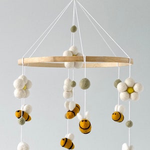 May include: A wooden baby mobile with felt bees, flowers, and balls. The bees are yellow and black, the flowers are white with yellow centers, and the balls are green.