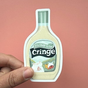 May include: A die-cut sticker in the shape of a salad dressing bottle. The label reads "It's Okay to be Cringe" with a cartoon cat eating pizza. The bottle is held by a hand against a pink background.