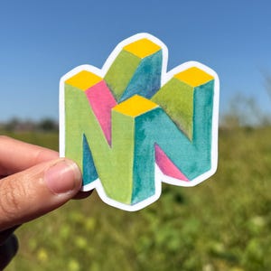 May include: A colorful sticker featuring the iconic Nintendo 64 logo. The logo is a stylized, three-dimensional 'N' shape, with green, pink, and teal coloring, and yellow accents. The sticker is held against a backdrop of a blue sky and green grass.