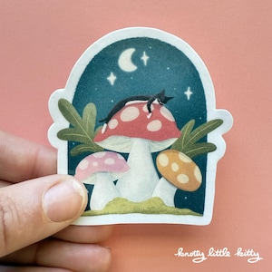 Könnte beinhalten: Eine schwarz-weiße Katze schläft auf einem rot-weißen Pilz mit zwei weiteren Pilzen, einem rosa-weißen und einem orange-gelben, vor einem Nachthimmel mit Sternen und einer Mondsichel.