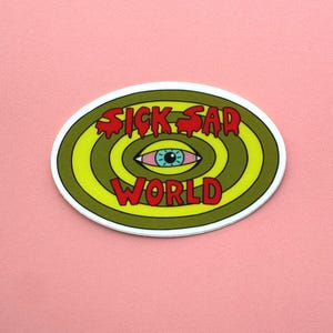 K&ouml;nnte beinhalten: Ovaler Aufkleber mit den Worten "SICK SAD WORLD" in roten, tropfenden Buchstaben. Das Design zeigt ein blau&auml;ugiges Auge in der Mitte, umgeben von konzentrischen gr&uuml;nen und gelben Kreisen, vor einem rosa Hintergrund.