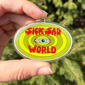 Könnte beinhalten: Ovaler Schlüsselanhänger mit dem Schriftzug "SICK SAD WORLD" in roter, stilisierter Schrift. Das Design zeigt ein zentrales Auge mit blauer Iris, umgeben von konzentrischen gelben und grünen Kreisen. Der Schlüsselanhänger hat ein klares, glänzendes Finish.