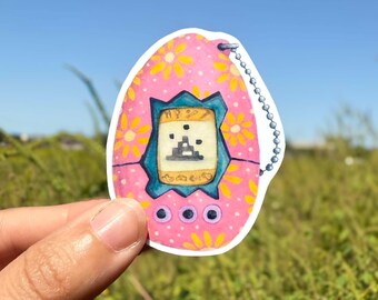 May include: A pink sticker in the shape of a retro handheld digital pet game. The sticker has a blue screen with a pixelated graphic and three pink buttons. Yellow flowers decorate the pink background. A silver chain is attached.