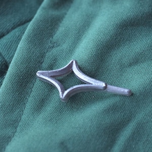 Puede incluir: Un pin de metal plateado en forma de estrella con un centro hueco. El pin está sobre un fondo de tela verde.