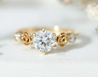 Könnte beinhalten: Ein goldener Ring mit einem zentralen, rund geschliffenen, klaren Edelstein in einer Krappenfassung. Die Ringschiene ist auf beiden Seiten des Mittelsteins mit skulpturalen Goldrosenmotiven verziert, flankiert von kleineren runden Akzentsteinen.