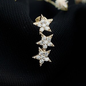 Può includere: Un orecchino color oro con tre ciondoli a forma di stella collegati in linea verticale. Ogni stella è incastonata con piccole pietre chiare e scintillanti. Il gioiello riporta la scritta WIIZLORY GEMS nell'angolo in alto a destra.