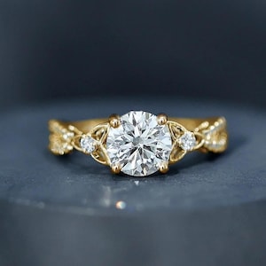 May include: A gold engagement ring with a large, round, clear gemstone at the centre. The ring features intricate filigree detailing and smaller accent stones. The ring is displayed on a dark grey surface. The text "WIIZLORY GEMS" is visible.