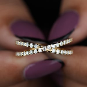 May include: A gold-coloured criss-cross ring, set with small, sparkling white gemstones. The ring is held between two hands with purple nail varnish. The brand name "WIIZLORY GEMS" is visible in the upper right corner.