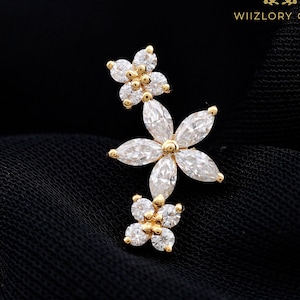 Könnte beinhalten: Ein goldfarbener Ohrring mit einem zentralen Blumendesign aus fünf klaren Steinen im Marquise-Schliff, flankiert von zwei kleineren vierblättrigen Blumenclustern aus runden klaren Steinen. Der Text WIIZLORY GEMS ist oben im Bild zu sehen.