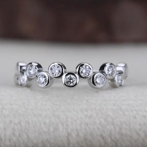 Könnte beinhalten: Ein silberner Ring mit einer Reihe kleiner, runder, klarer Edelsteine, die entlang des Bandes gesetzt sind. Die Edelsteine sind gleichmäßig verteilt und bilden ein zartes, funkelndes Design. Der Ring wird auf einer weichen, cremefarbenen Oberfläche präsentiert.