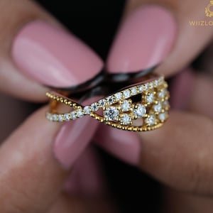 May include: A gold ring with a crisscross design, encrusted with small, sparkling diamonds. The ring features a cluster of diamonds in the center and is held by a person with pink nail polish. The ring is a piece of jewelry.