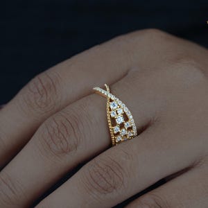May include: A gold-colored ring with a unique crisscross design, adorned with multiple small, clear gemstones. The ring is set on a hand with a dark background, showcasing the intricate details and sparkle of the jewelry. The ring is from WIIZLORY GEMS.