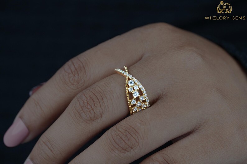 May include: A gold-colored ring with a unique crisscross design, adorned with multiple small, clear gemstones. The ring is set on a hand with a dark background, showcasing the intricate details and sparkle of the jewelry. The ring is from WIIZLORY GEMS.