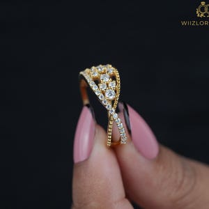 May include: A gold-colored ring with a crisscross design, adorned with numerous small, clear gemstones. The ring features a cluster of larger stones at the top. The background is black, and the ring is held by a person with pink nail polish. The text "WIIZLORY GEMS" is visible.