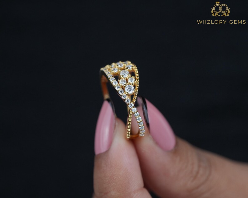 May include: A gold-colored ring with a crisscross design, adorned with numerous small, clear gemstones. The ring features a cluster of larger stones at the top. The background is black, and the ring is held by a person with pink nail polish. The text "WIIZLORY GEMS" is visible.