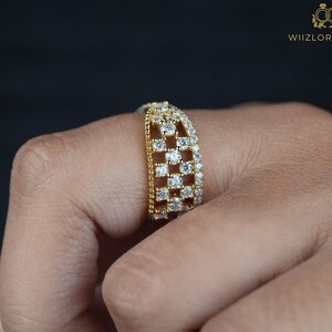 May include: A gold-toned ring with a grid-like design, encrusted with numerous small, sparkling white gemstones. The ring is worn on a finger, showcasing its intricate details and the brilliance of the stones. The ring is from WIIZLORY GEMS.