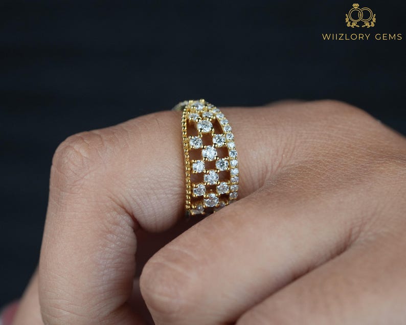 May include: A gold-toned ring with a grid-like design, encrusted with numerous small, sparkling white gemstones. The ring is worn on a finger, showcasing its intricate details and the brilliance of the stones. The ring is from WIIZLORY GEMS.