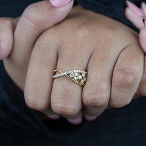 May include: A gold-toned ring with an intertwined design, adorned with small, sparkling gemstones. The ring features a delicate, interwoven pattern with a central cluster of stones, creating a sophisticated and elegant look. The ring is displayed on a hand.