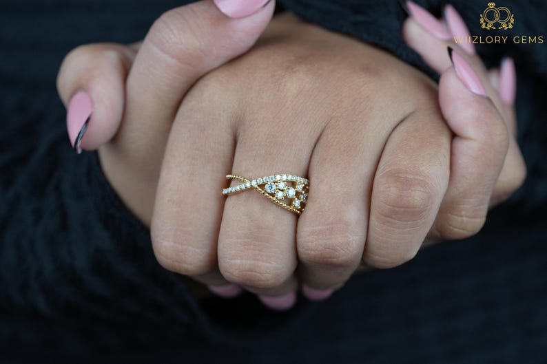 May include: A gold-toned ring with an intertwined design, adorned with small, sparkling gemstones. The ring features a delicate, interwoven pattern with a central cluster of stones, creating a sophisticated and elegant look. The ring is displayed on a hand.