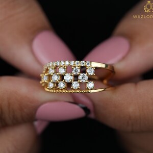 May include: A gold ring with a grid design, set with multiple small, sparkling diamonds. The ring is held against a dark background, showcasing its intricate details and the brilliance of the gemstones. The ring is a piece of jewelry.