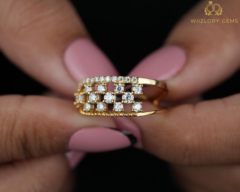 May include: A gold ring with a grid design, set with multiple small, sparkling diamonds. The ring is held against a dark background, showcasing its intricate details and the brilliance of the gemstones. The ring is a piece of jewelry.
