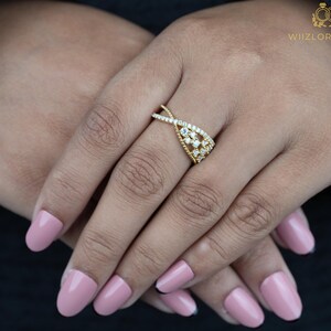 May include: A gold-colored ring with a unique design featuring small, clear gemstones. The ring has a twisted band and a central cluster of stones. The ring is on a person's hand with pink nail polish.