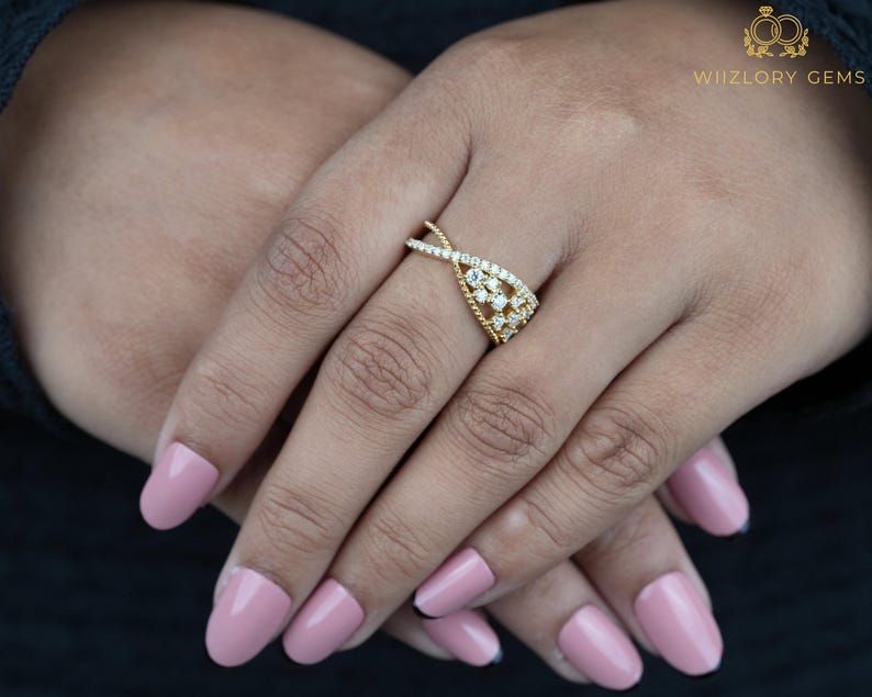 May include: A gold-colored ring with a unique design featuring small, clear gemstones. The ring has a twisted band and a central cluster of stones. The ring is on a person's hand with pink nail polish.