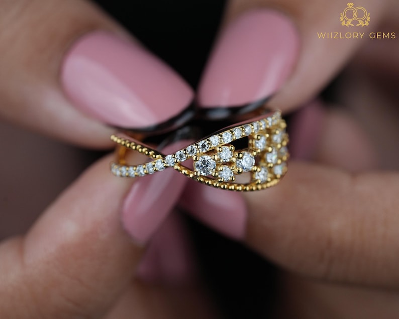 May include: A gold ring with a crisscross design, encrusted with small, sparkling diamonds. The ring features a cluster of diamonds in the center and is held by a person with pink nail polish. The ring is a piece of jewelry.