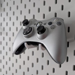 May include: A silver video game controller with black joysticks and buttons is mounted on a white pegboard. The controller is held in place by a white bracket. The pegboard has a pattern of evenly spaced, oblong holes.