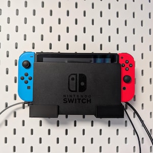 May include: A black Nintendo Switch console with blue and red Joy-Con controllers is mounted on a white pegboard. The console is centered, with the text "NINTENDO SWITCH" visible. Cables extend from the console.