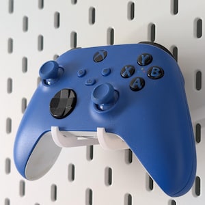 May include: A blue games controller with black buttons and joysticks is mounted on a white pegboard. The controller is held in place by a white bracket. The pegboard has evenly spaced holes.