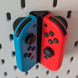 May include: A pair of Nintendo Switch Joy-Con controllers, one blue and one red, mounted on a white pegboard. The controllers are connected and show the button layout and analog sticks.