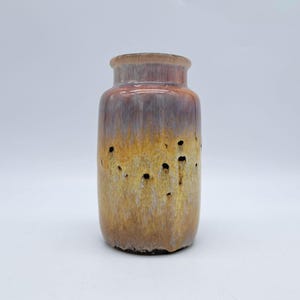 Puede incluir: Jarrón de cerámica cilíndrico con borde acampanado. El jarrón presenta un degradado de color, pasando de un marrón rojizo en la parte superior a un amarillo dorado en la inferior. Tiene pequeños agujeros oscuros en el cuerpo.