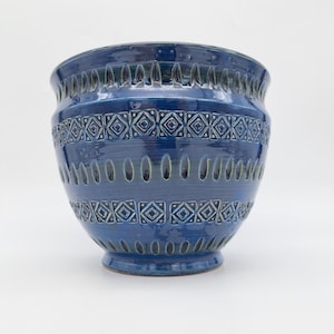Peut inclure: Un pot de fleurs en céramique bleu profond avec un corps large et arrondi et un bord évasé. L'extérieur est orné de bandes horizontales de motifs géométriques en losange en relief et d'indentations ovales verticales, avec un fini brillant.