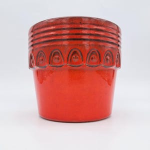 May include: A vibrant red ceramic plant pot featuring a smooth, glossy finish on the lower section and a textured upper rim. The rim is decorated with a series of dark, recessed triangular arches and horizontal grooves encircling the top.