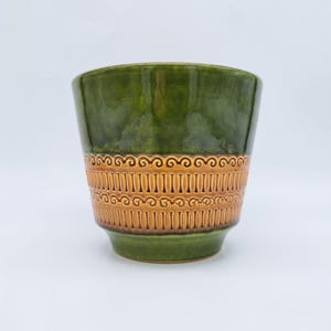 Puede incluir: Una maceta de cerámica verde con una banda de detalles marrones texturizados. La maceta tiene un acabado brillante y una forma cilíndrica, adecuada para plantas de interior. El diseño presenta un patrón repetitivo de líneas verticales y espirales.