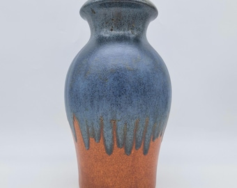 Könnte beinhalten: Eine Keramikvase mit blau-brauner Glasur. Die Vase hat einen abgerundeten Körper und einen ausgestellten Rand. Die blaue Glasur tropft an den Seiten herunter und geht in einen braunen Sockel über. Die Vase ist etwa 20 cm hoch.