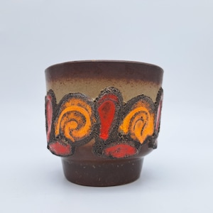 May include: A ceramic planter with a brown base and rim, featuring a tan middle section. The planter is adorned with a band of orange and red swirl designs. The design is raised and textured.
