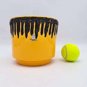 Puede incluir: Una maceta de cerámica amarilla con un diseño de pintura negra goteando alrededor del borde. Un pequeño emblema está adherido al costado. Una pelota de tenis amarilla está al lado de la maceta.