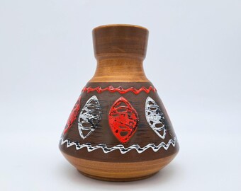 May include: A brown ceramic vase with a wide base and a narrow neck. The vase features a decorative pattern of red, white, and black leaf-shaped designs. A white zigzag pattern borders the design. The vase is approximately 7 inches tall.