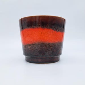 Puede incluir: Una maceta de cerámica con forma cilíndrica. La maceta presenta un degradado de colores, con una parte superior marrón oscuro, una banda naranja vibrante en el medio y una base marrón más oscura. Adecuada para uso en interiores o exteriores.