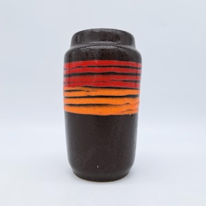 Puede incluir: Un jarrón de cerámica marrón oscuro con bandas horizontales rojas y naranjas. El jarrón tiene forma cilíndrica con una parte superior ligeramente ensanchada. Las bandas de color crean un efecto de puesta de sol. El jarrón es probablemente vintage o retro.
