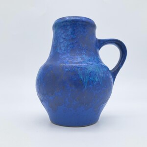 May include: A ceramic pitcher featuring a bulbous body and a single handle. The surface is finished in a vibrant cobalt blue glaze with mottled, darker blue and black textured patterns that create a layered, variegated appearance across the exterior.