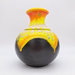 Könnte beinhalten: Eine Keramikvase mit bauchigem Körper und engem Hals. Die untere Hälfte ist in mattem Schwarz gehalten, während der obere Bereich ein lebhaftes Tropfglasur-Muster in leuchtendem Gelb, Orange und Weiß aufweist, das einen Effekt erzeugt.