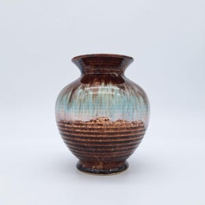 Könnte beinhalten: Eine Keramikvase mit bauchigem Körper und ausgestelltem Rand. Die Vase hat eine braune Oberseite, einen hellblauen und braunen Mittelteil und einen dunkelbraunen, gerippten Sockel. Die Vase ist etwa 20 cm hoch.