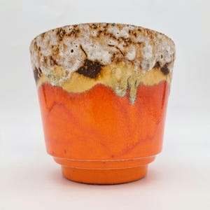 Op de afbeelding: Een keramische beker met een feloranje basis die overgaat in een gele band, bekroond met een getextureerde, kratervormige rand in bruin- en wittinten. De beker heeft een getrapte, taps toelopende voet en een gladde, glanzende afwerking onderaan.