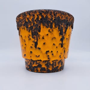 Peut inclure: Un pot de fleurs en céramique avec un corps émaillé orange vif et un motif texturé en forme de coulures brun foncé rappelant de la lave, partant du bord et couvrant la base. Le récipient a une forme cylindrique conique avec une arête horizontale.