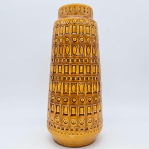 Puede incluir: Un jarrón alto de cerámica en tonos marrón y dorado. El jarrón presenta un patrón repetitivo de rectángulos y círculos, creando una superficie texturizada. El diseño es simétrico, con una base ancha y una parte superior más estrecha.